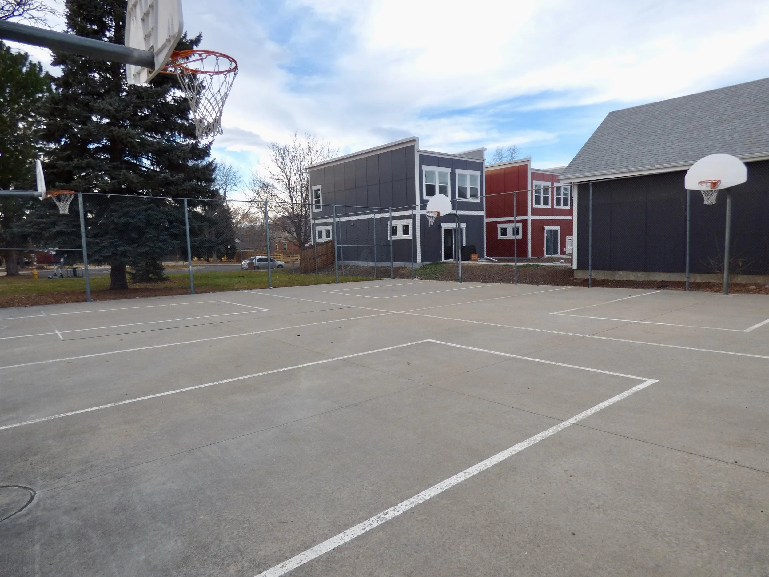 An outdoor basketball court surrounded by a chain-link fence, with two basketball hoops, residential houses, and trees in the background under a partly cloudy sky at Madame C.J. Walker Park, Denver, CO 80205.