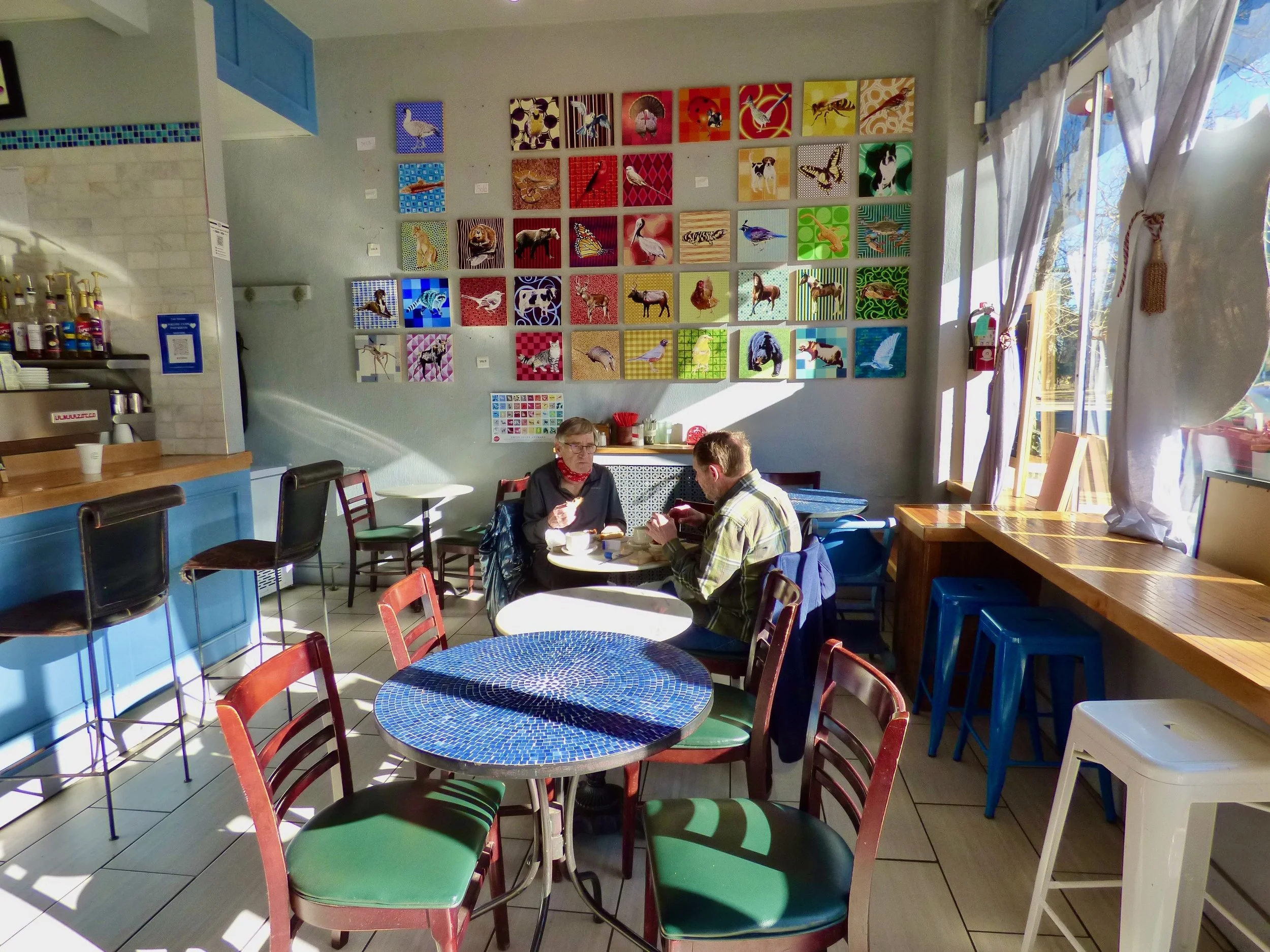 Interior of a cafe with colorful artwork on the wall, two people sitting at a table, sunlight coming through large windows at Cafe Miriam, Denver, CO 80205.