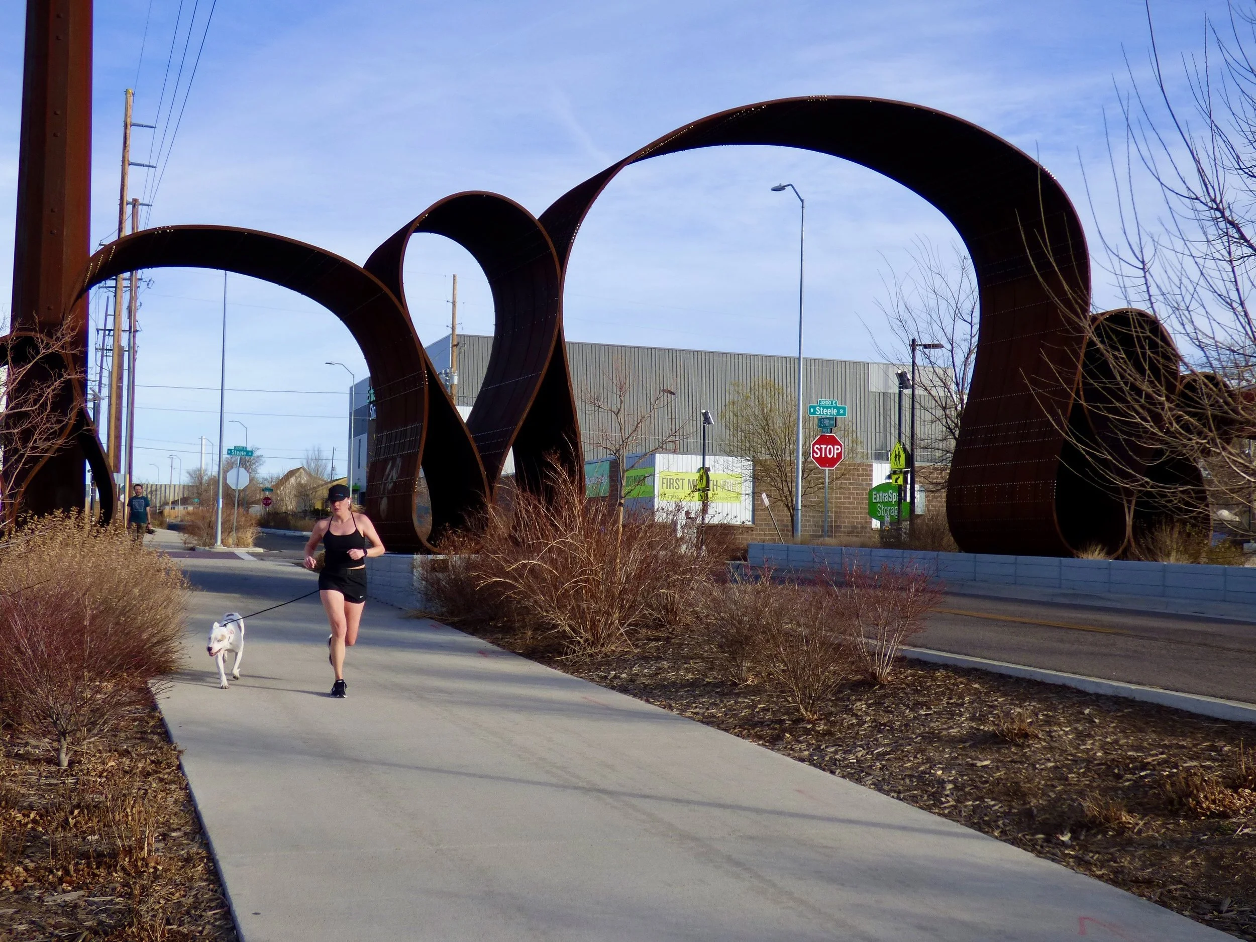 A woman jogging a dog on a sidewalk with a large metal sculpture of intertwined rings and street signs visible in the background in 39th Ave Greenway, Denver, CO 80205.
