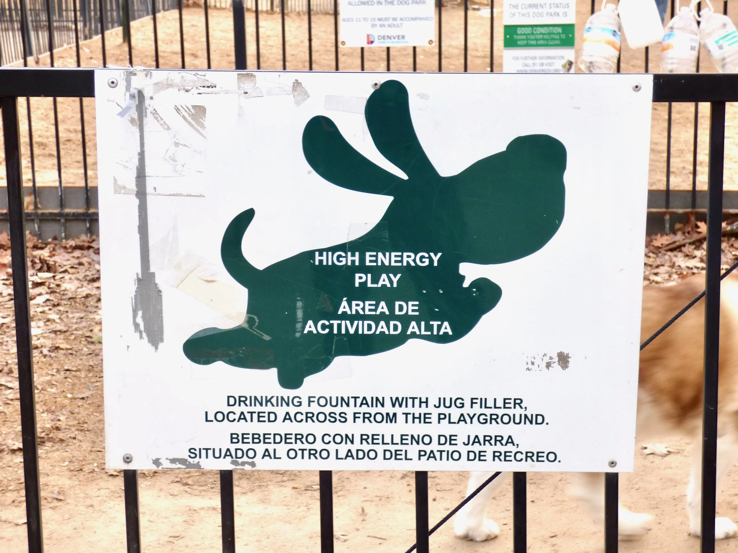Sign at a dog park indicating a high energy play area with a silhouette of a dog and a butterfly. Text in English and Spanish about drinking fountain with jug filler.