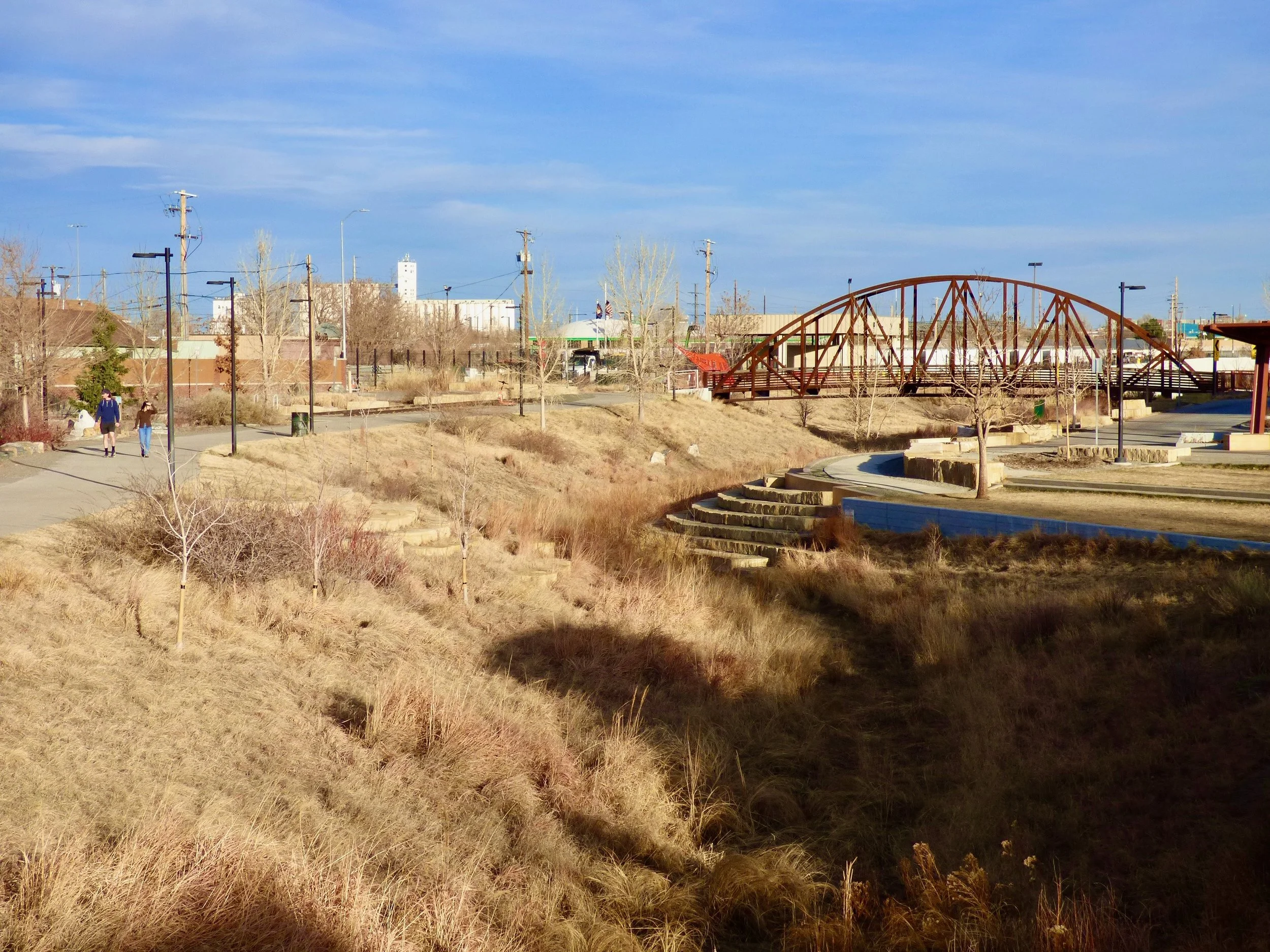 A park with walking paths, leafless trees, a small brown hill, and a red bridge in the distance. Two people are walking on the path on the left side of the image. The sky is partly cloudy  in 39th Ave Greenway, Denver, CO 80205.