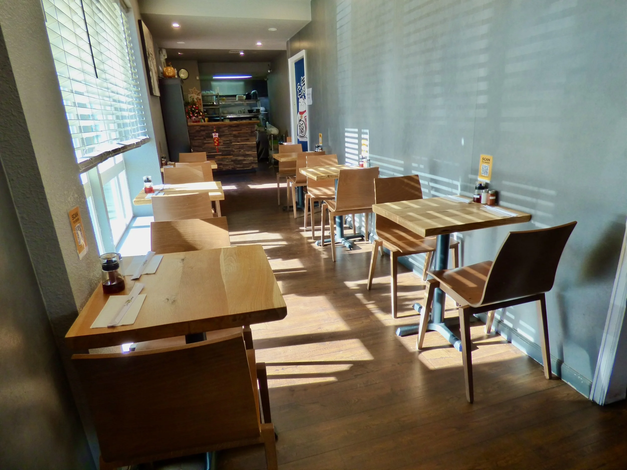 Empty small restaurant or cafe with wooden tables and chairs, sunlight streaming through window blinds, QR code sign on table, condiments on tables, and a counter in the background.