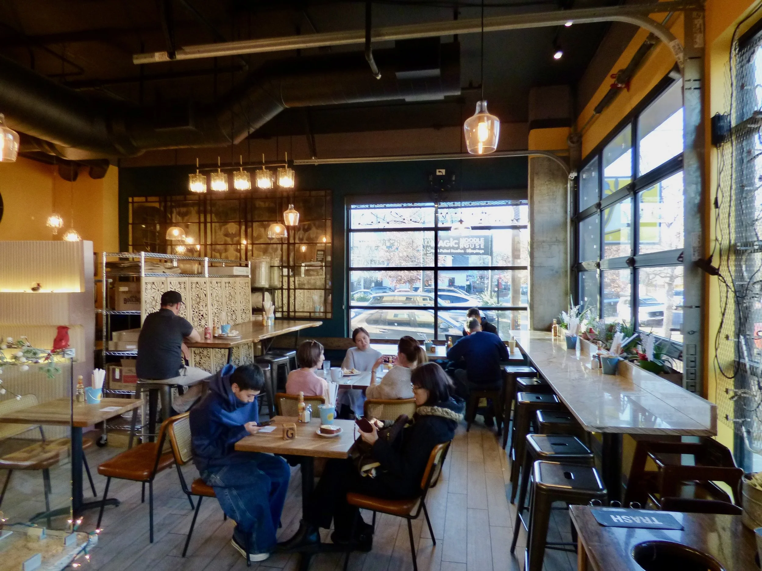 Interior of a cozy cafe with large windows, wooden tables, and pendant lighting. Several people are sitting at tables and the counter, some using phones and others eating at Olive & Finch, Denver, CO.