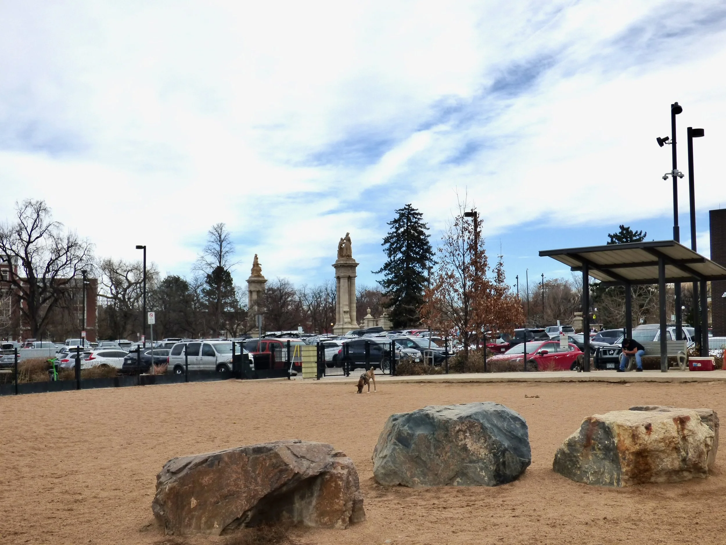 A dog sitting on the sand of a park with large rocks in the foreground, a parking lot with many cars, trees, and architectural columns in the background, and a man sitting under a small roof shelter.