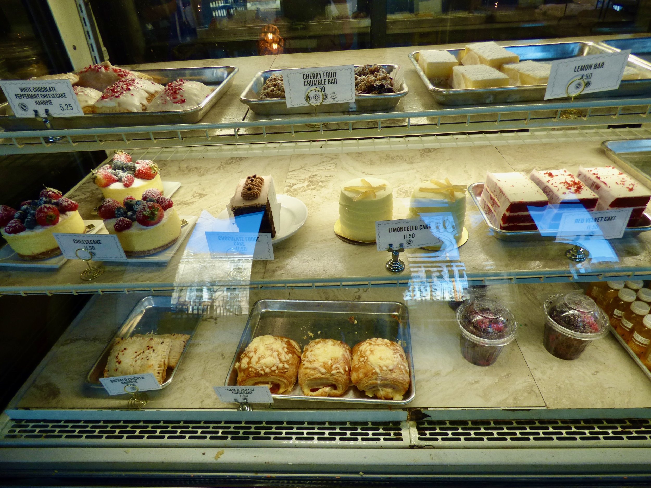 Display case with various desserts, including cheesecakes topped with berries, chocolate fudge cake, lemon cake, red velvet cake, and pastries, with handwritten price labels at Olive & Finch, Denver, CO.