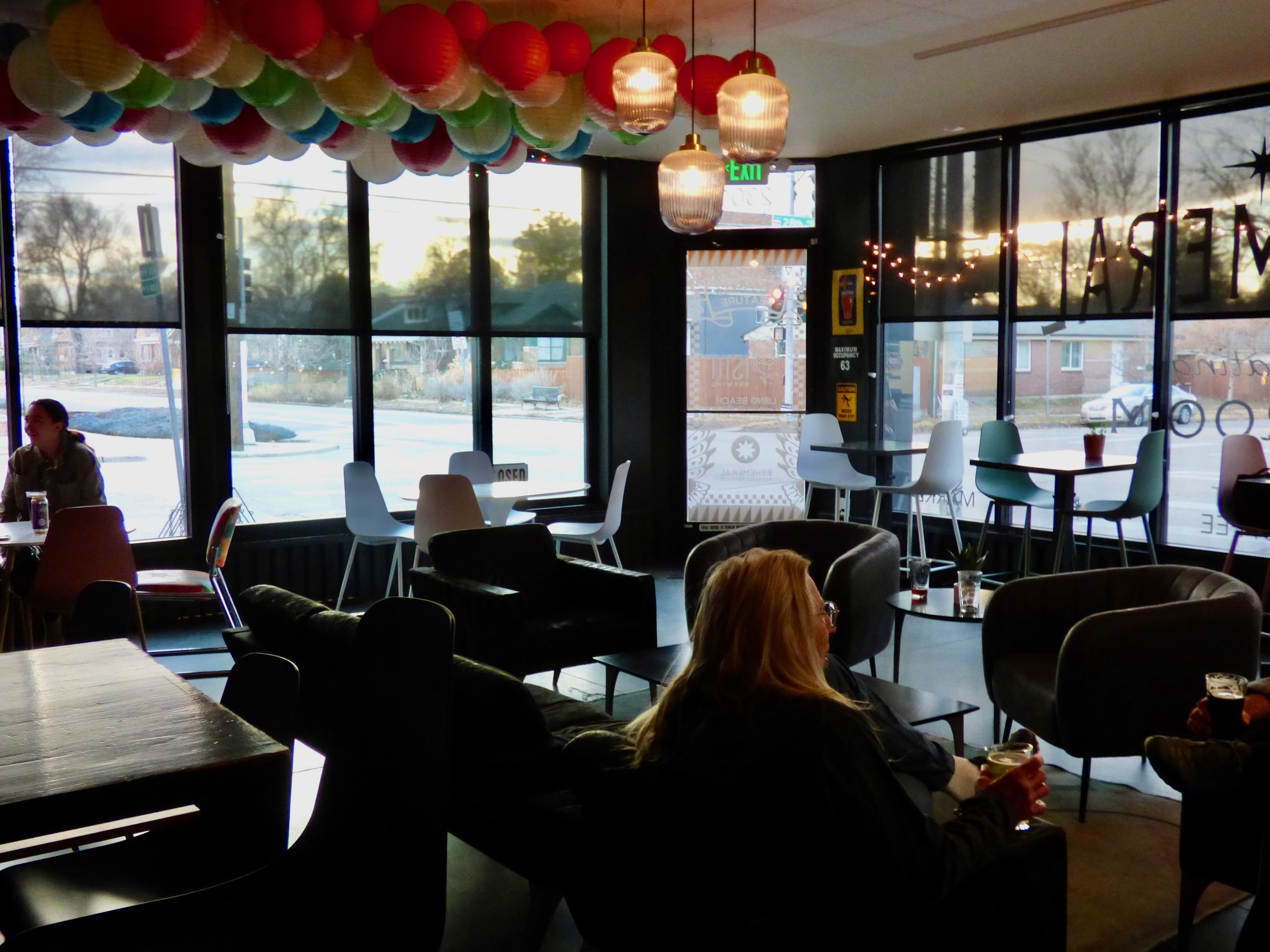 Interior of a cafe with large windows showing a snowy scene outside, decorated with colorful hanging paper lanterns and string lights, with people sitting at tables and sofas at Ephemeral Rotating Taproom, Denver, CO 80205.