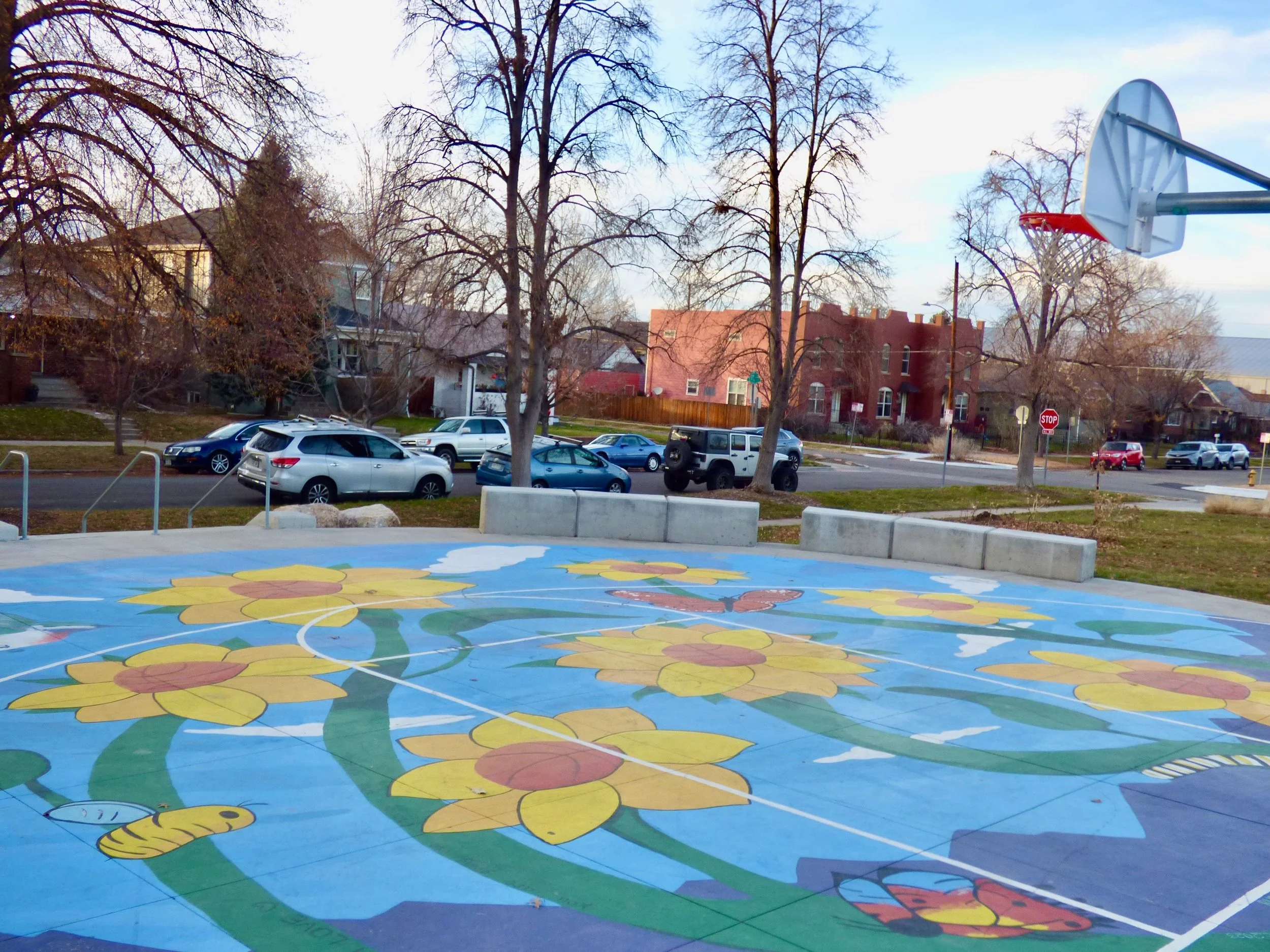 A colorful outdoor basketball court with a painted floral design, surrounded by trees and parked cars, with houses in the background and a basketball hoop overhead in Russell Square Park, Denver, CO 80205.