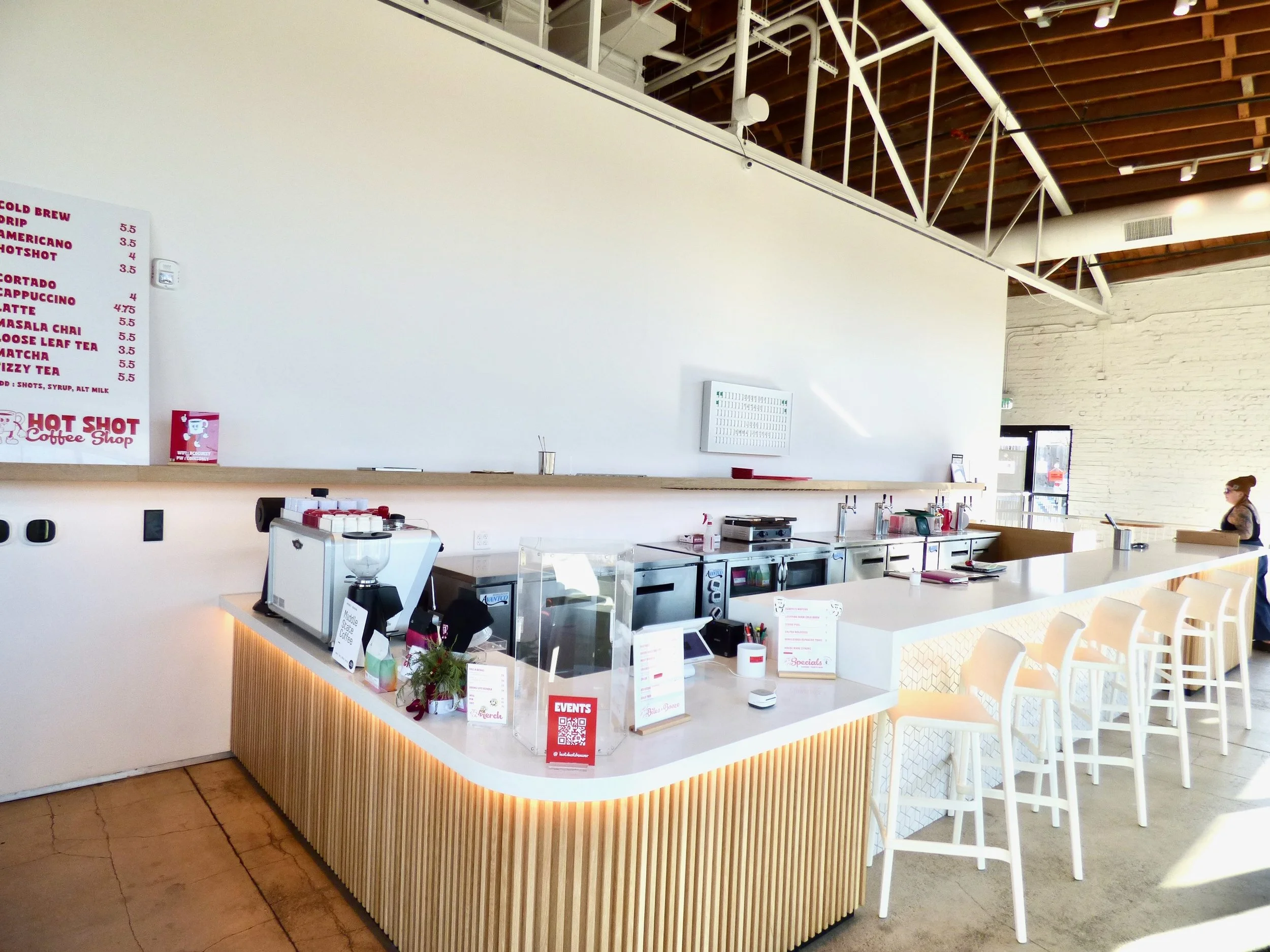 Interior of a modern coffee shop with a white counter, coffee machines, and bar stools, with a woman on the right side near a door, and a menu sign on the left wall displaying drink options and prices at Hot Shot Coffee Shop, Denver, CO 80205.