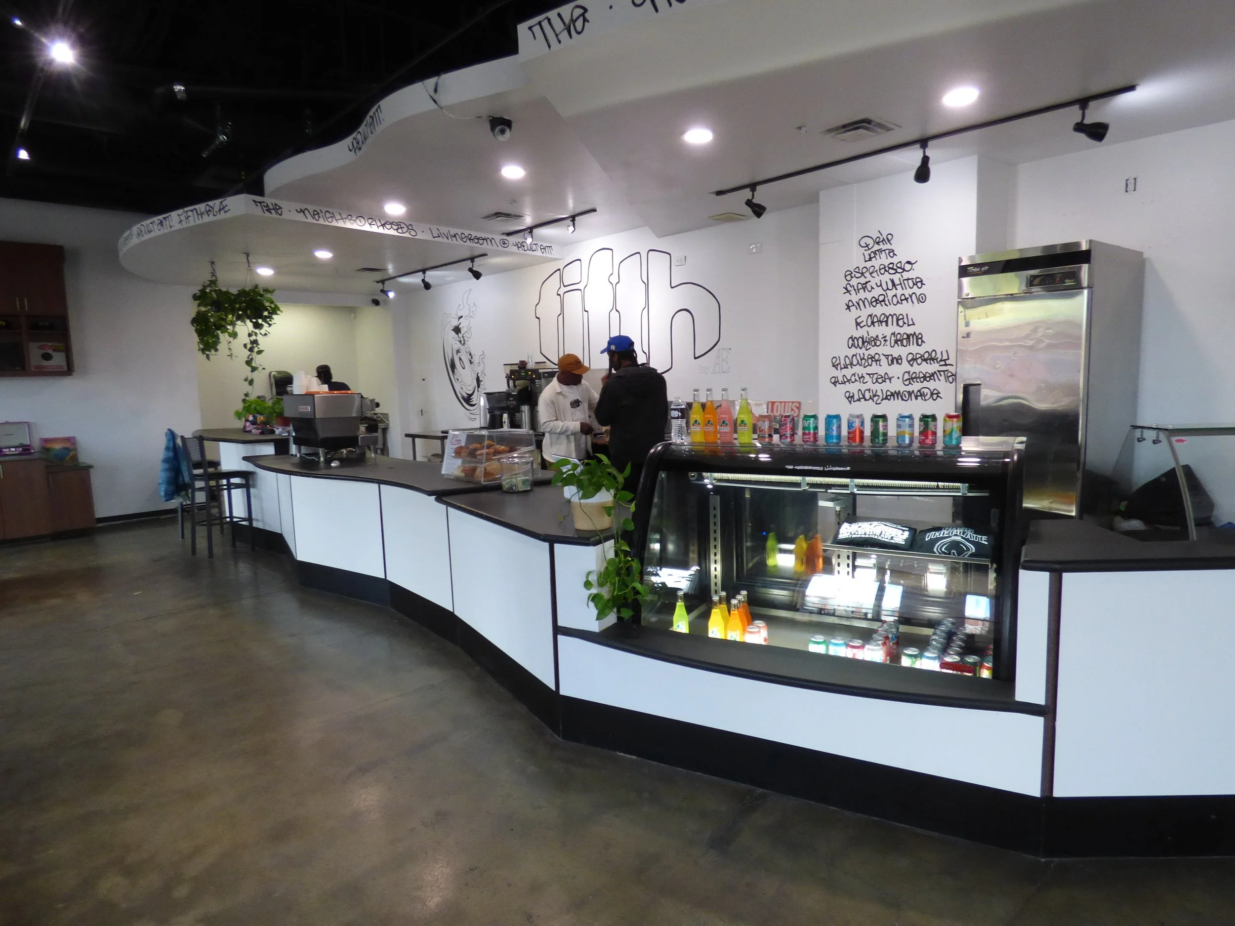 Interior of a modern cafe or pastry shop with a curved counter, coffee machines, and a small refrigerator. There are two staff members and some decorative plants, with menu items written on a white wall at Fifth Coffee House, Denver, CO 80205.