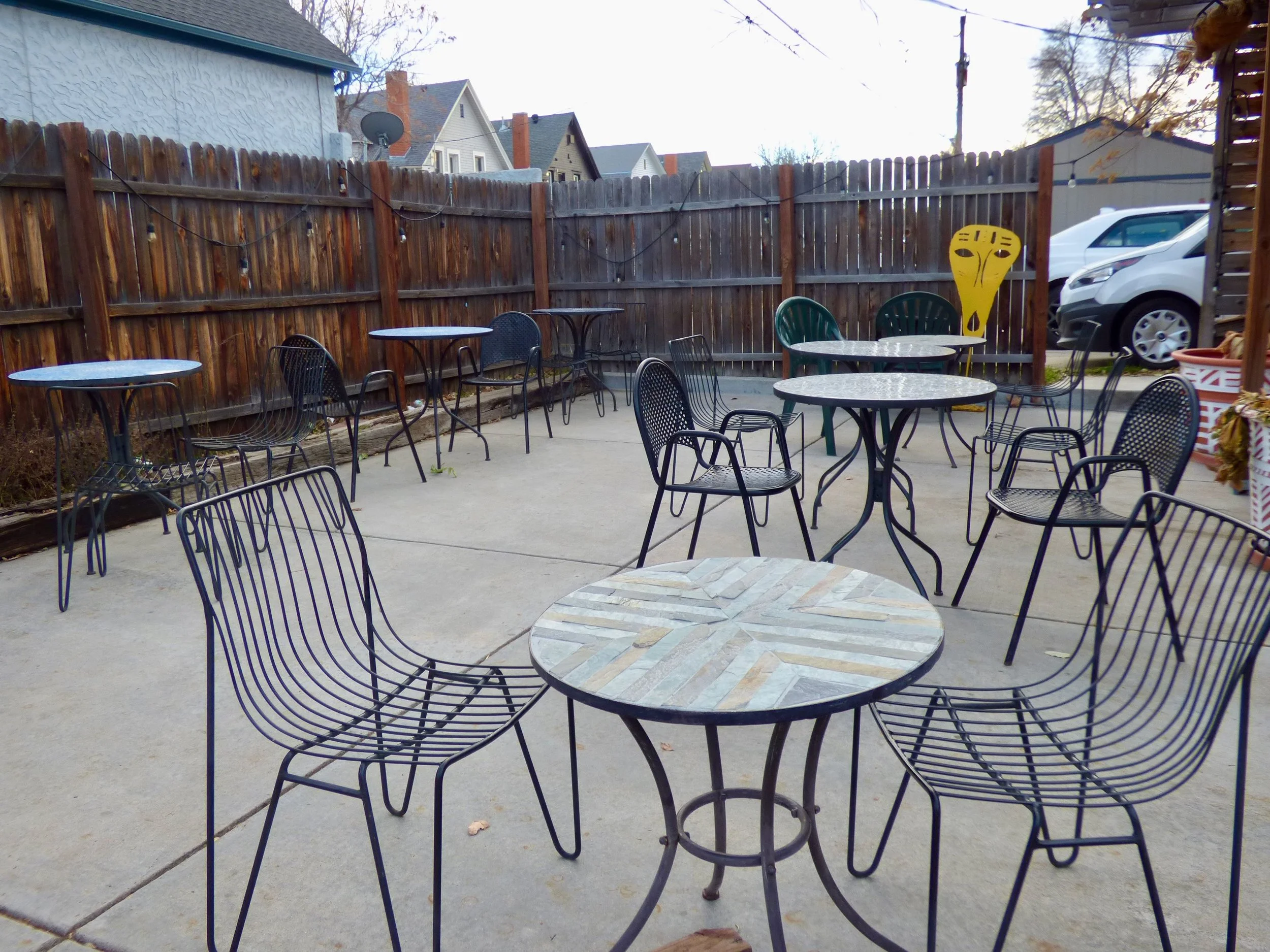 Outdoor patio with metal tables and chairs, wooden privacy fence, some string lights, parked cars, and a decorative yellow sculpture at Whittier Cafe, Denver, CO 80205.
