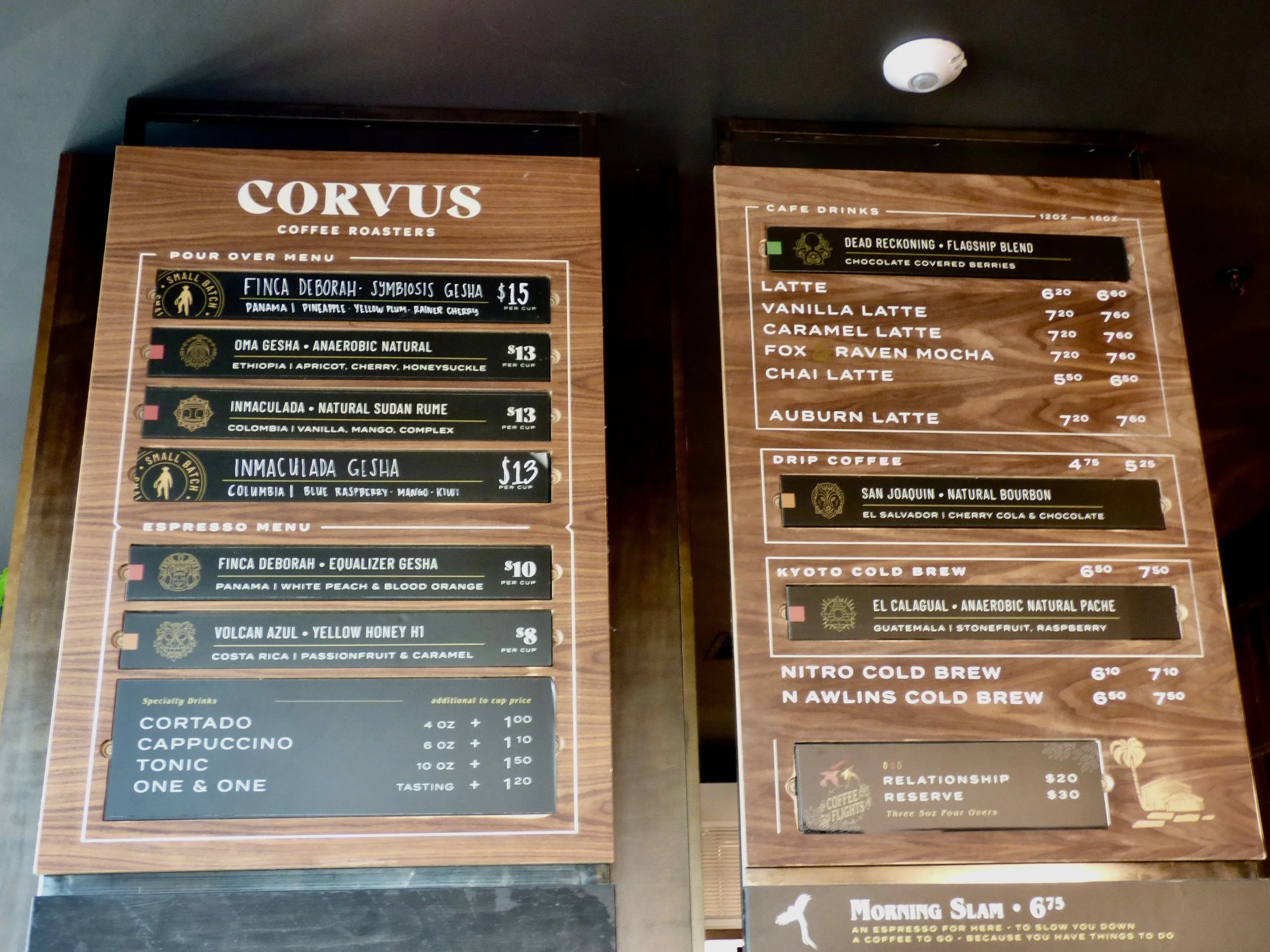 Menu boards inside a coffee shop displaying various coffee drinks, prices, and specialty beverages at Corvus Coffee Roasters, Denver, CO 80205.