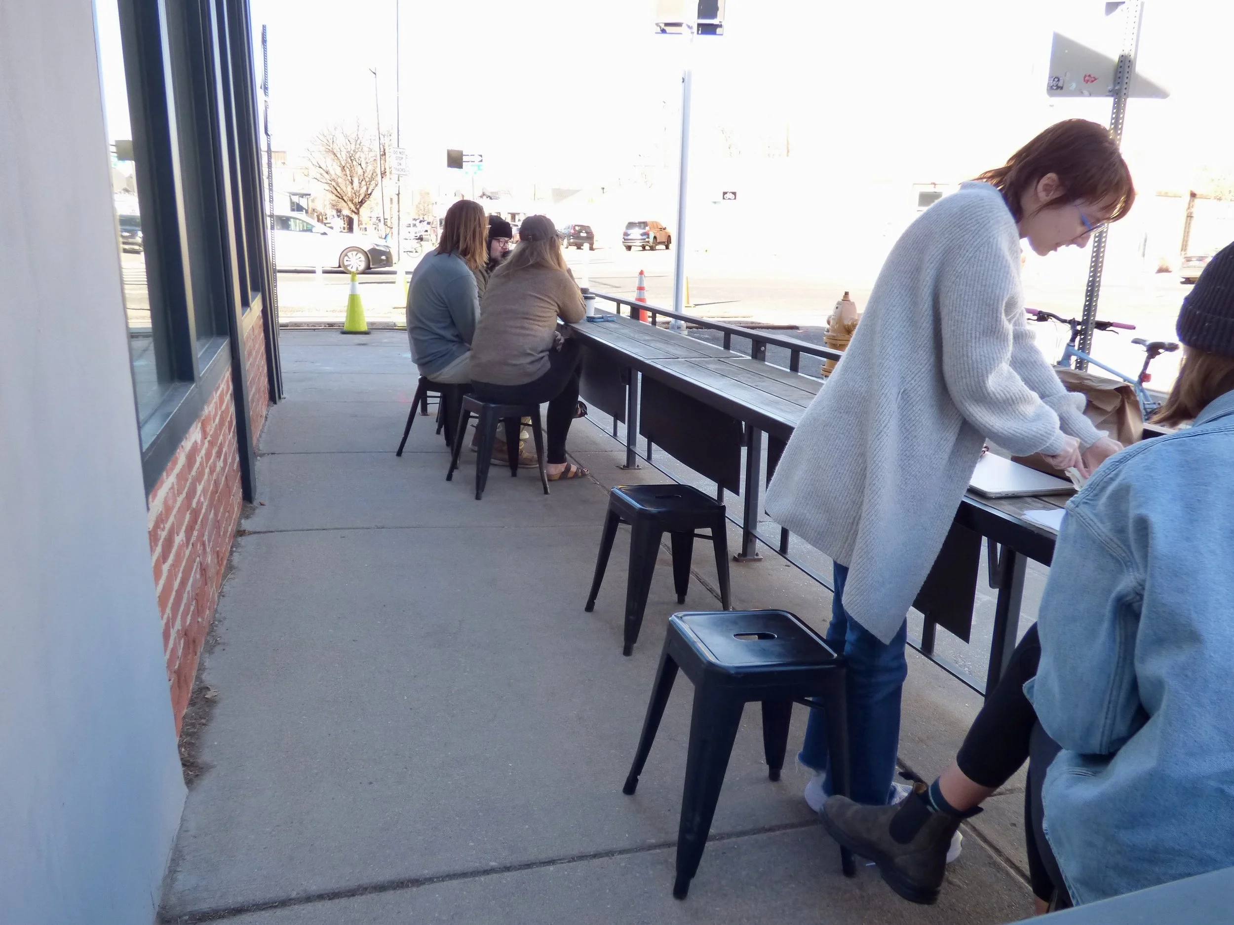 People seated outside at a cafe or restaurant on a sidewalk, with some individuals working on laptops and others talking, during daytime at Queen City Collective Coffee House, Denver, CO 80205.