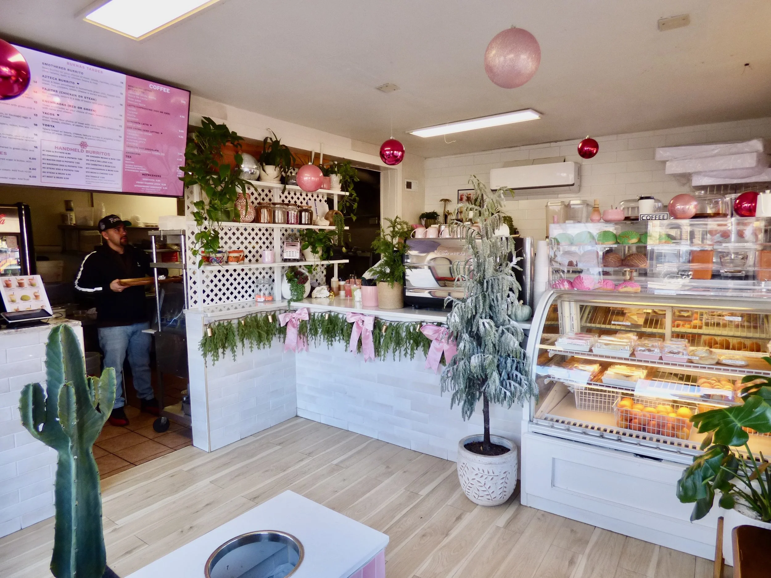 Interior of a bakery or cafe decorated with pink and red ornaments, potted plants, a white counter with pink ribbons, a display case with baked goods, and a menu board on the wall at Oasis Cafe & Grill, Denver, CO 80205.