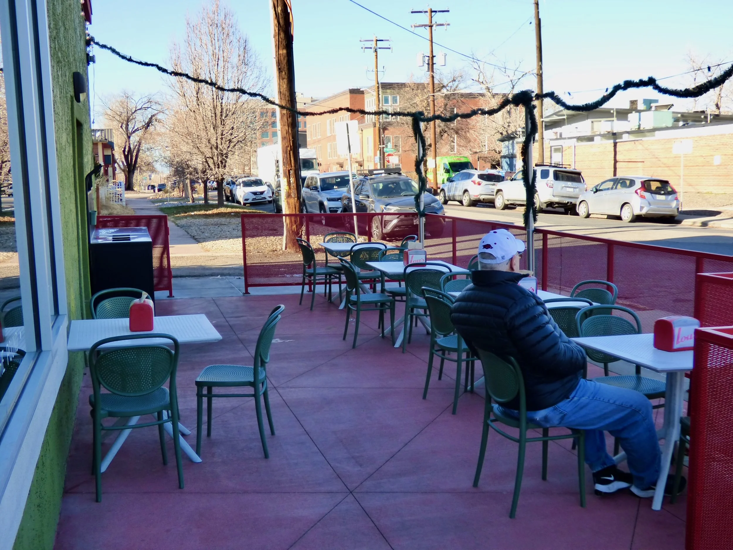 Empty outdoor patio of a restaurant or café with green chairs and tables, and one person wearing a black jacket and white cap sitting alone. The patio is enclosed with red railing, and the background shows a street with parked cars and leafless trees.