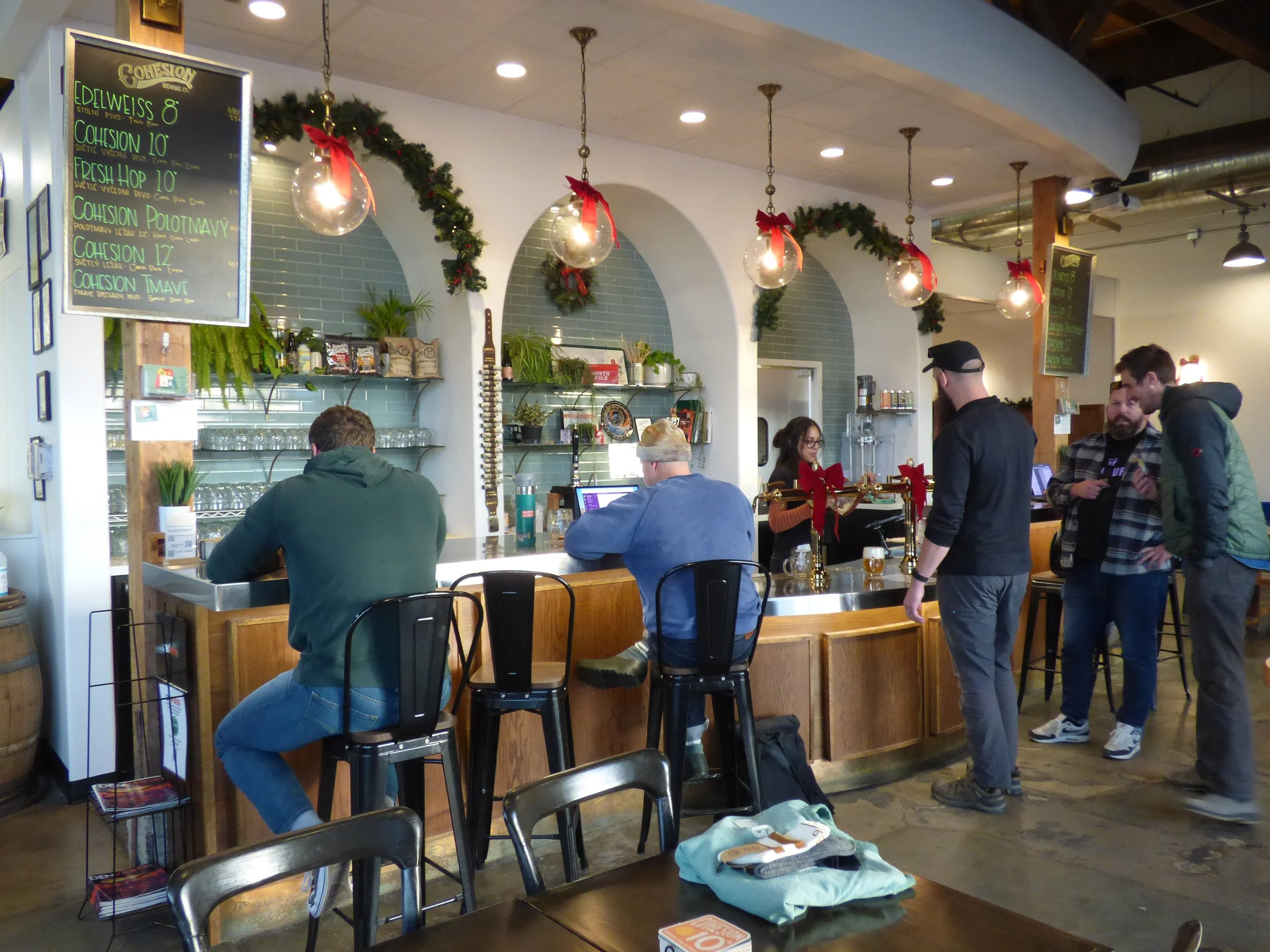 Inside a bar decorated with Christmas garlands and ornaments, with customers ordering drinks and a bartender working at Cohesion Brewery, Denver, CO 80205.