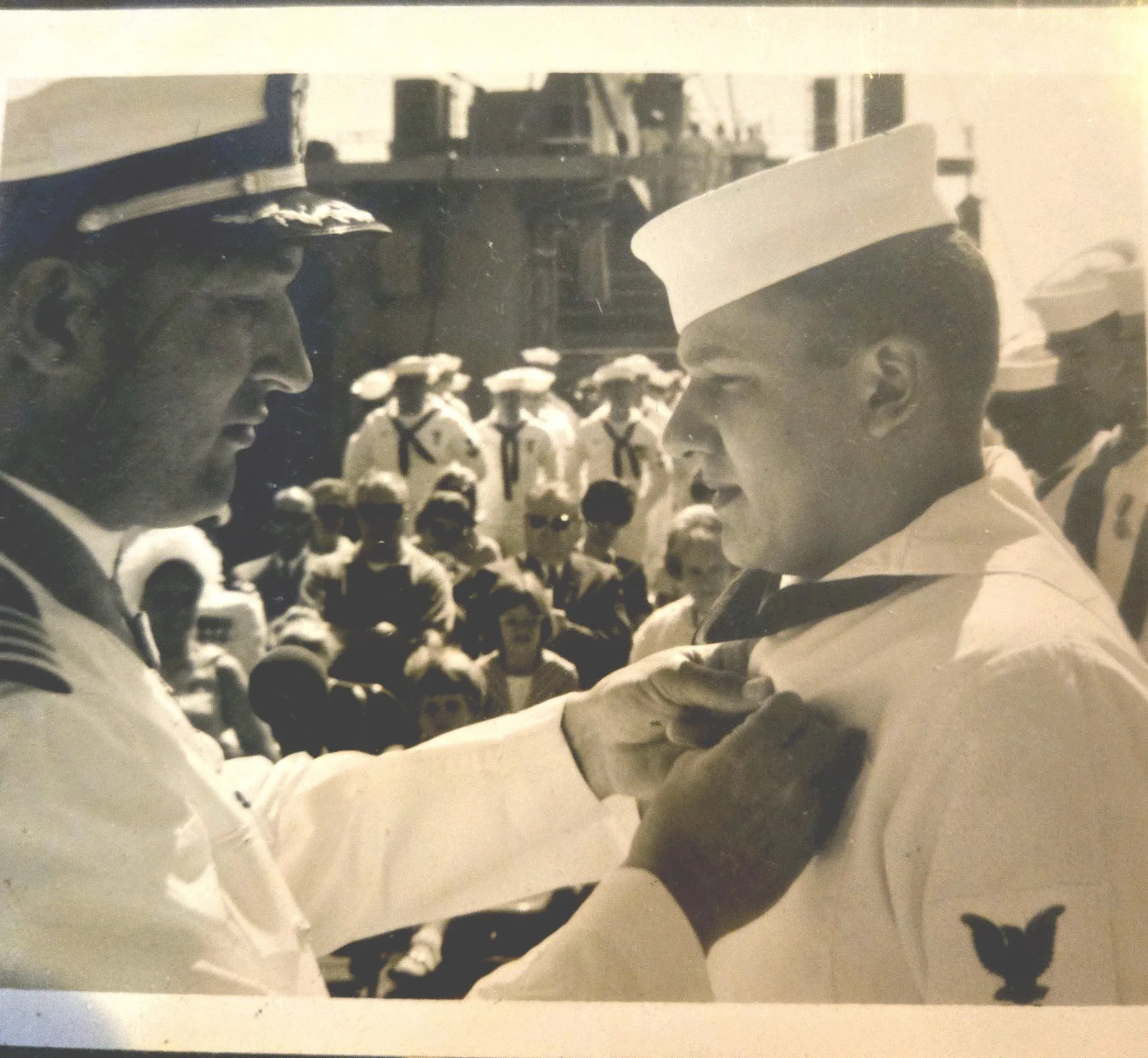 Black and white photograph of two sailors in uniform exchanging a handshake and a salute during a military ceremony, with a crowd of sailors in the background.