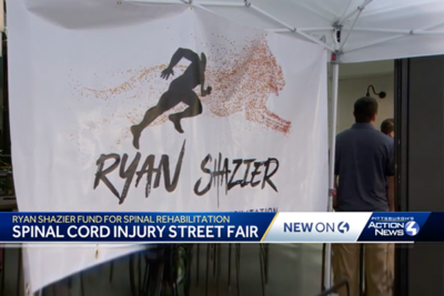 A banner displaying a silhouette of a person running with the name Ryan Shazier, promoting a fund for spinal rehabilitation at a street fair. A white tent with people is in the background.