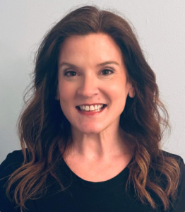 A woman with wavy brown hair smiling at the camera, wearing a black top against a plain light background.