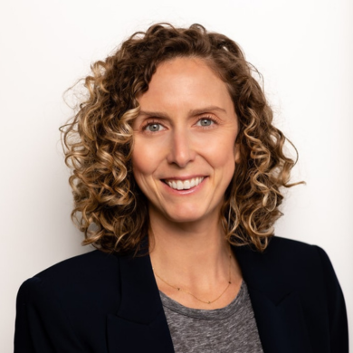 Headshot of a woman with curly blond hair, smiling, wearing a dark blazer and a gray top, against a white background.