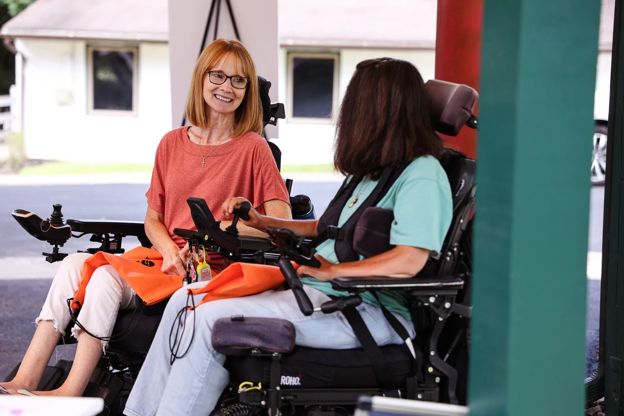 Two women with disabilities in wheelchairs having a conversation, one smiling and the other holding a remote control.