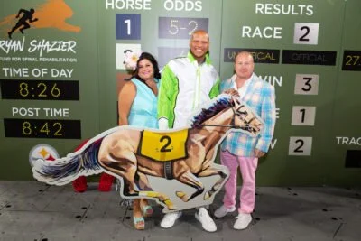 Three people standing next to a large racehorse cutout with the number 2, in front of a green scoreboard showing race results and odds