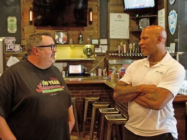 Two men having a conversation in a bar or restaurant, with drinks and bar equipment visible in the background.