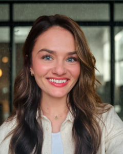 Close-up of a woman with wavy brown hair and blue eyes smiling at the camera, wearing a white shirt, with a dark, blurry indoor background.