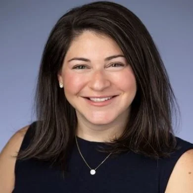 Portrait of a smiling woman with shoulder-length dark hair, wearing a sleeveless black top and a necklace, against a blue background.