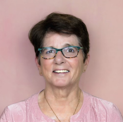 A smiling older woman with short dark hair, wearing glasses, a pink top, and small earrings, standing against a pink background.