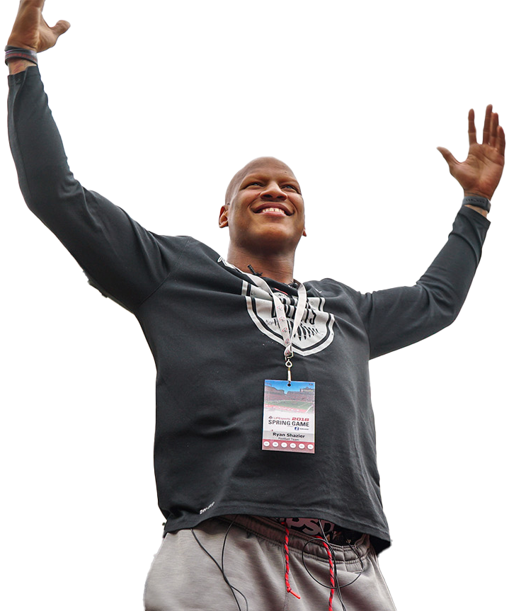 A man smiling and raising his arms, wearing a black hoodie with a logo, an accreditation badge hanging around his neck, and gray sweatpants, at an outdoor event.