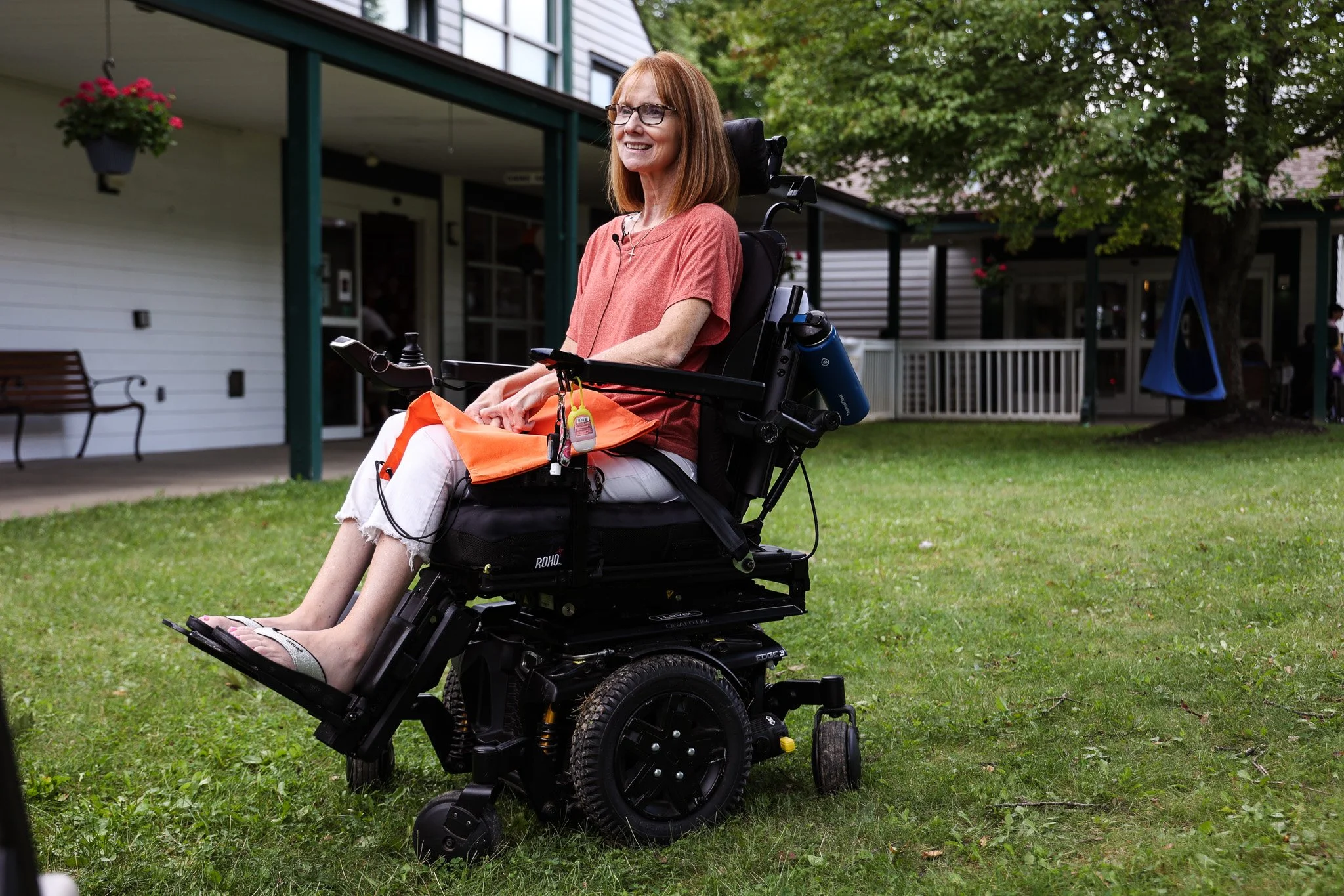 A woman with glasses and shoulder-length red hair sitting in a motorized wheelchair outdoors on a grassy area, smiling. She is wearing a coral-colored top and white pants. In the background, there is a building with a porch, and trees with green leaves.