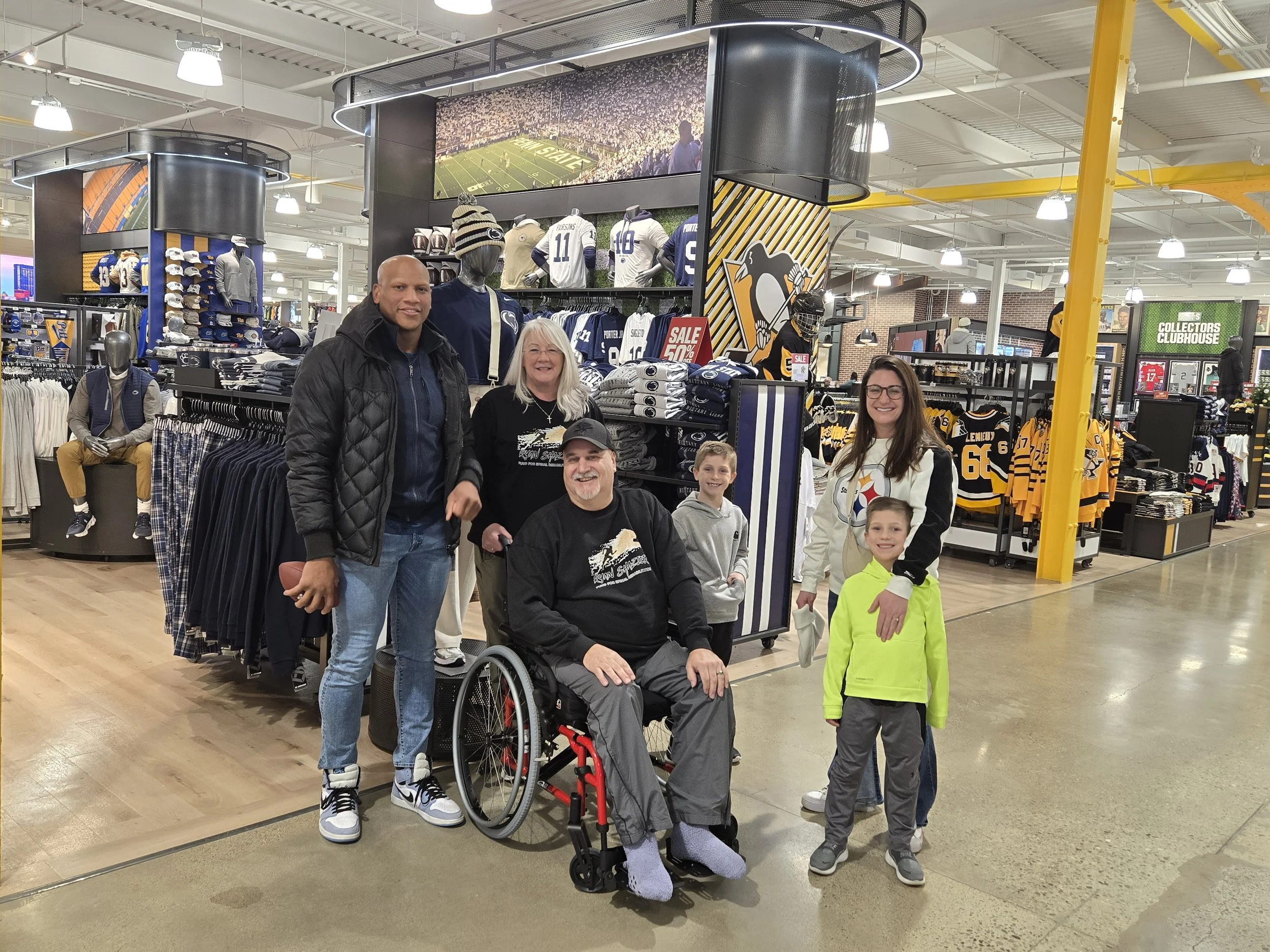 A group of six people, including a man in a wheelchair, poses inside a sports apparel store featuring Pittsburgh Steelers merchandise. The background shows shelves with jerseys, hats, and other team gear.