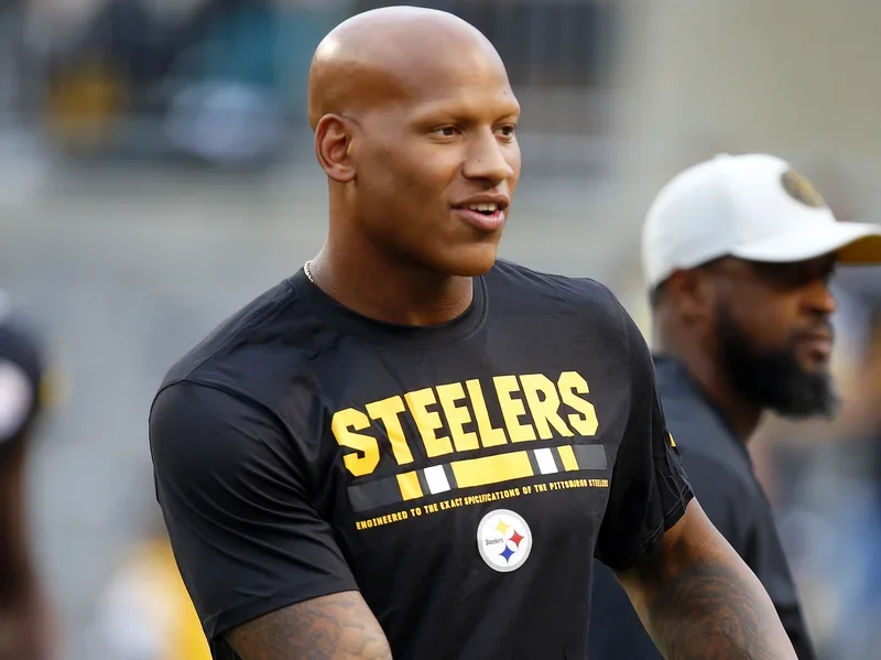 A man in a black T-shirt with the yellow word 'Steelers' and a Steelers logo, standing outdoors with other people.