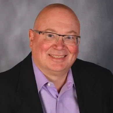 A bald man wearing glasses, smiling, dressed in a purple shirt and black blazer, against a gray background.