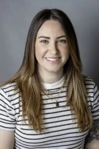 A young woman with long brown hair, smiling, wearing a white and black striped shirt and a gold necklace.
