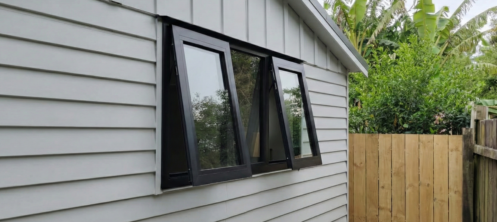 A house with gray horizontal siding and a black-framed, fold-out window that is open. The window is partially folded outward, and there are green trees and a wooden fence in the background.
