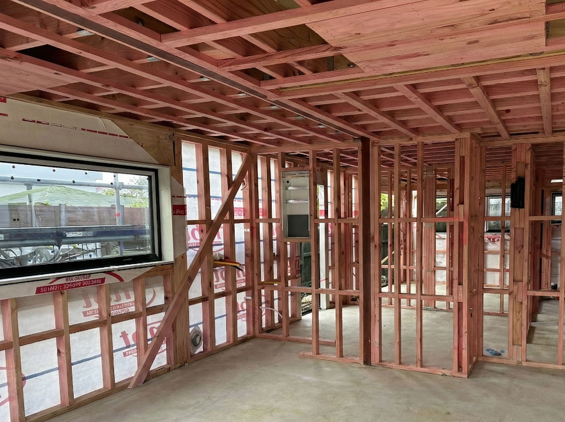 The interior of a house under construction, with exposed wooden framing for walls and ceiling, a window with a view outside, and a concrete floor.