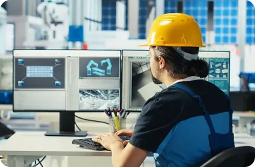 A construction worker wearing a yellow helmet working at a desk with multiple computer monitors showing building designs and blueprints.