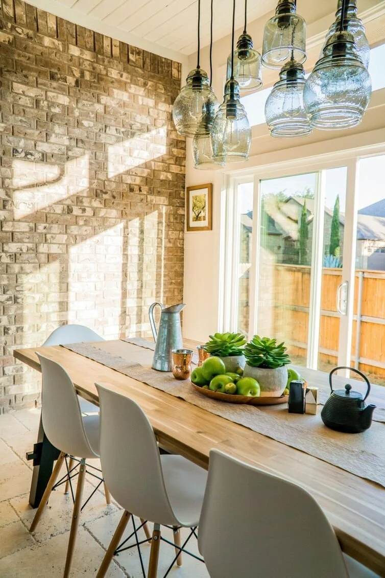 Bright dining room with a wooden table, white chairs, a brick wall, and a large window with a view of the backyard. Hanging glass jar pendant lights are above the table, and decorative succulents and apples are on the table.