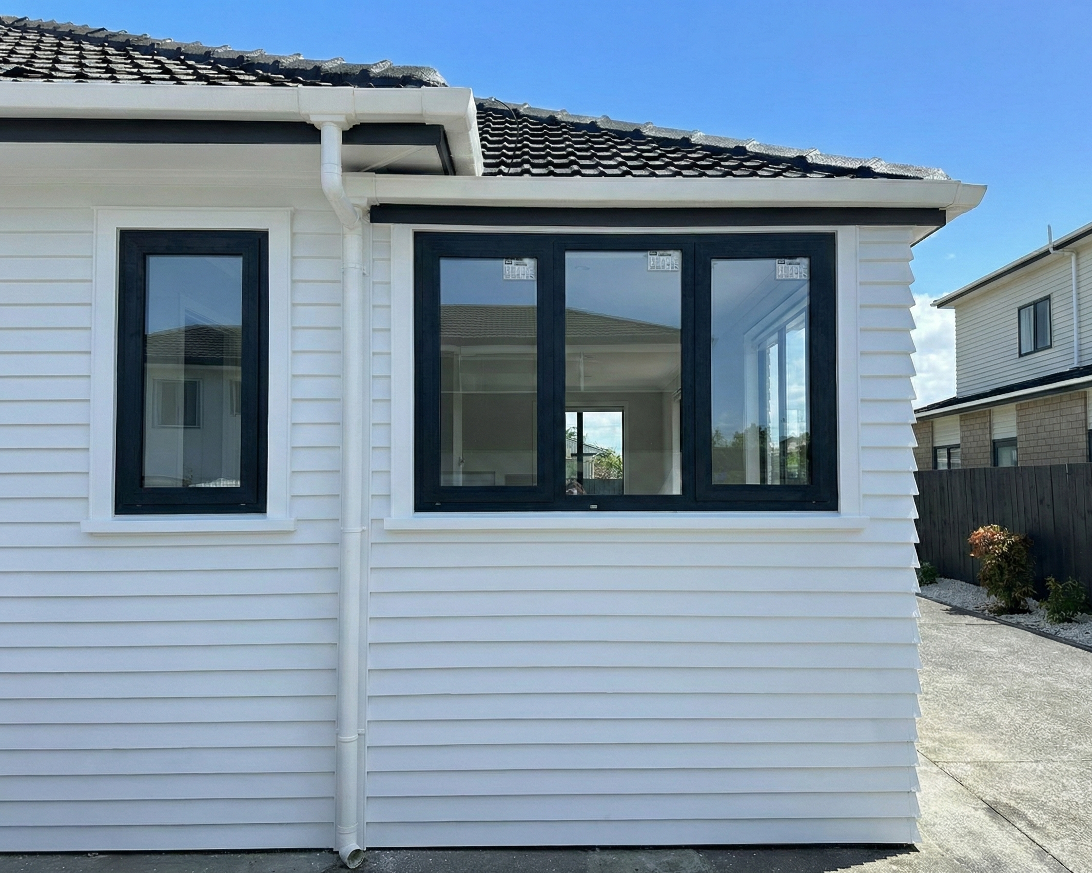 The exterior of a white house with black-framed windows and a partial view of a dark roof, with a concrete driveway and a small garden area on the right under a clear blue sky.