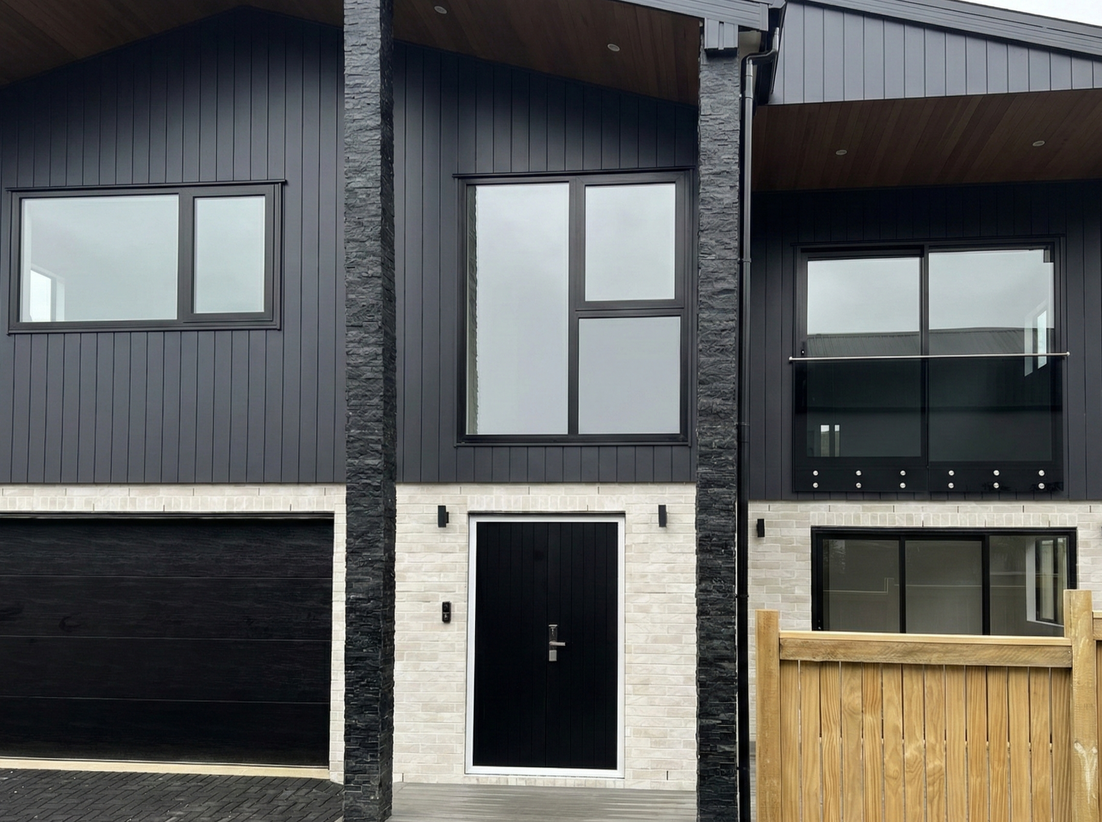 Modern multi-story house with black siding, white brick accents, large windows, a black front door, a garage with black door, and a wooden fence.