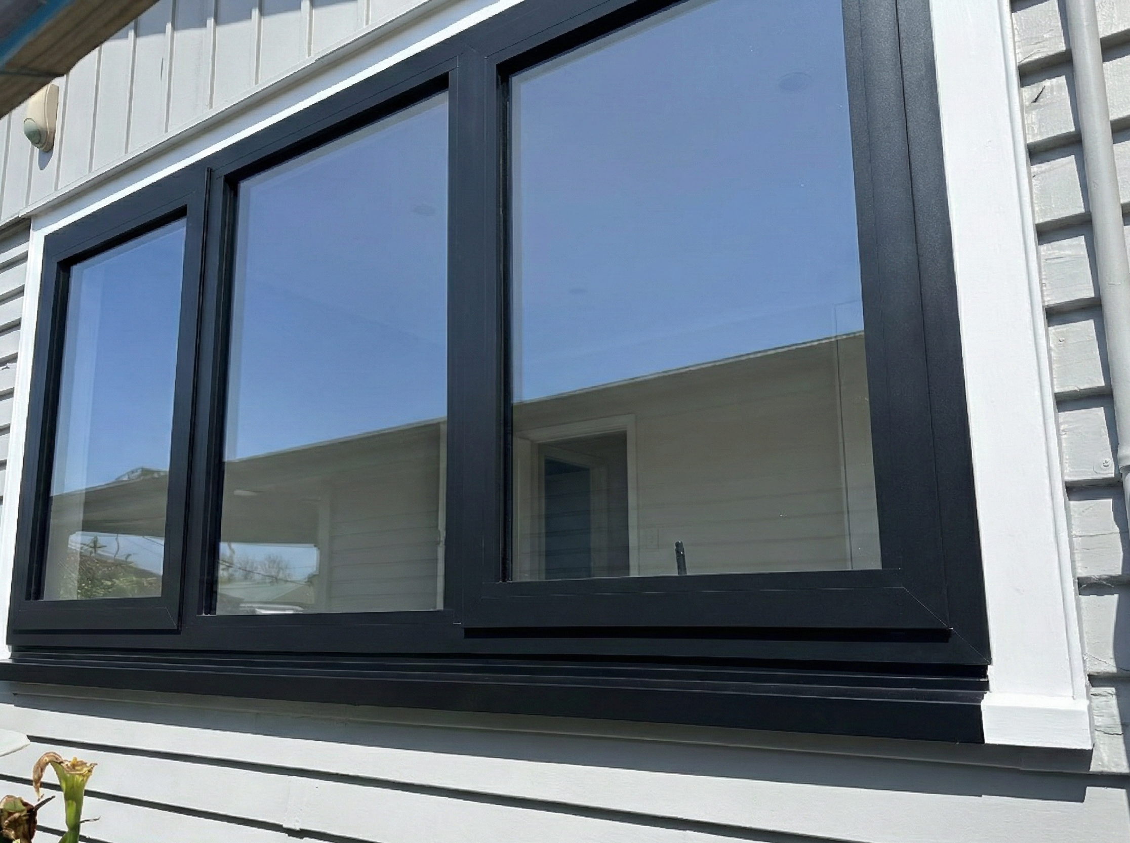 Close-up of a window with black frame on the side of a house with white siding, reflecting a blue sky and part of the roof.