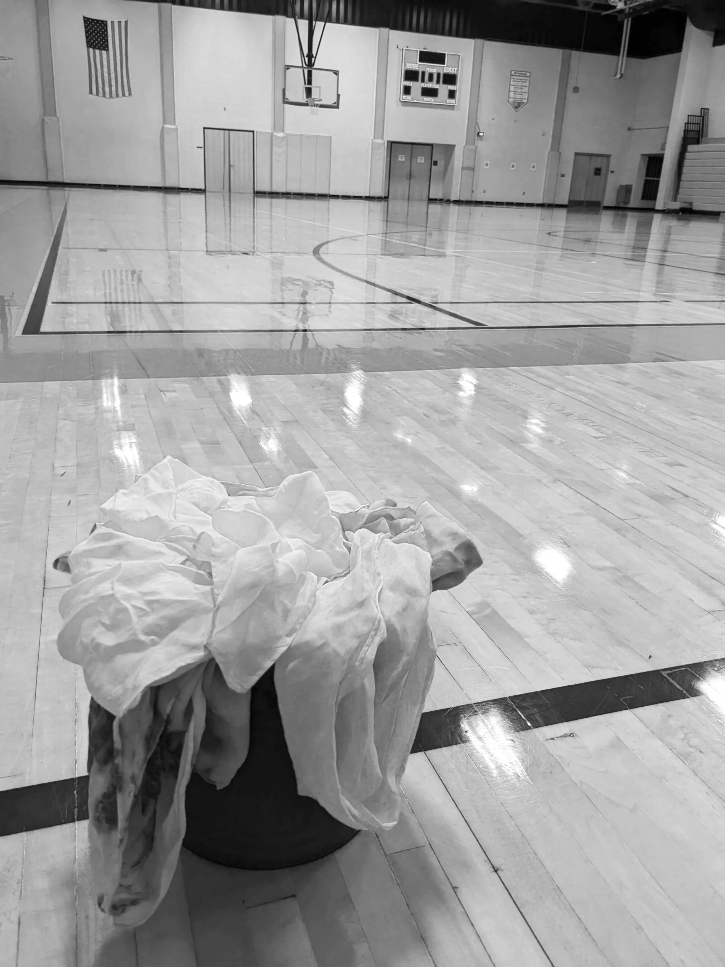 An empty basketball gymnasium with a wrinkled white cloth in the foreground and an American flag on the wall. The gym has polished wooden floors, a basketball hoop, and a scoreboard above. The area appears to be prepared for an event or maintenance.