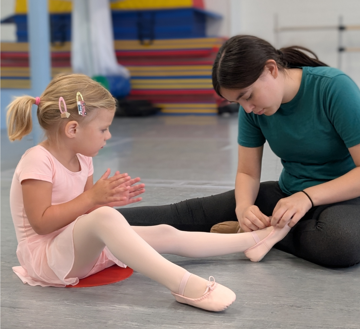 A young girl in pink ballet attire receiving a foot stretch from her ballet instructor in a dance studio.
