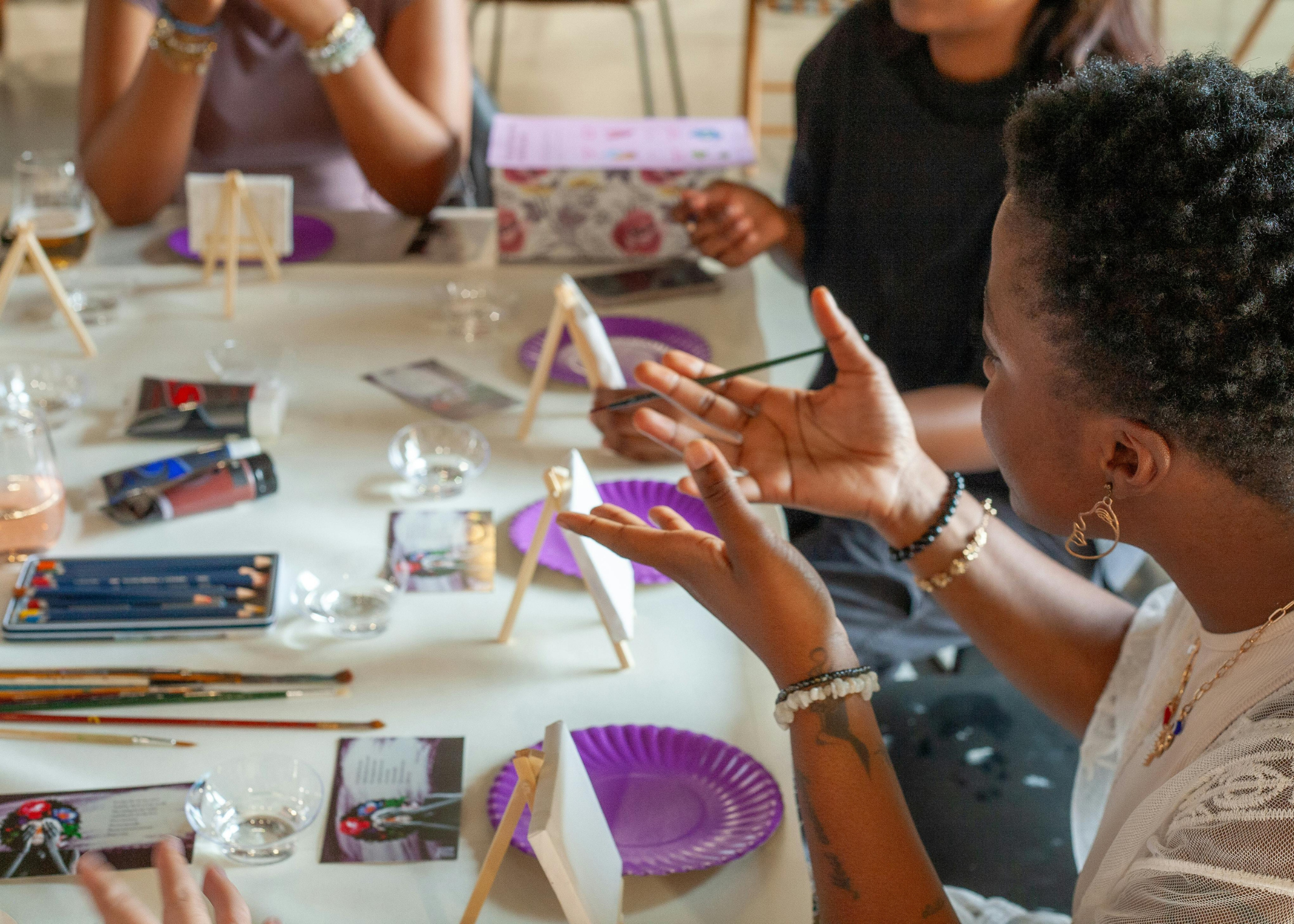 People sitting around a table at an art class, with paintbrushes, water glasses, and small easels. A woman in the foreground is holding a paintbrush and explaining something, wearing jewelry and a white top.