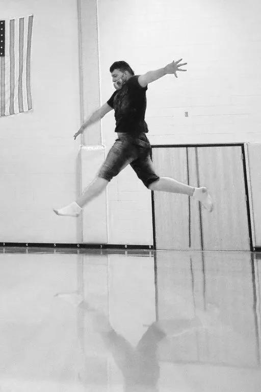 Person jumping mid-air indoors, with a wooden wall and an American flag in the background.