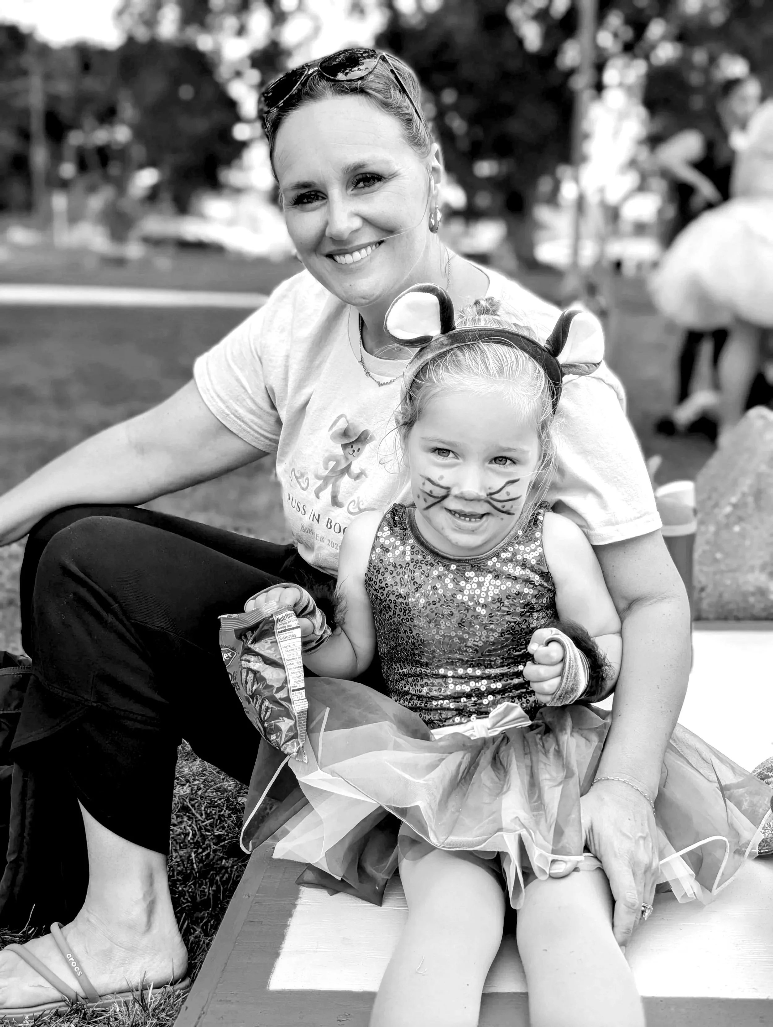 A smiling woman and young girl at an outdoor event. The woman has sunglasses on her head and the girl has face paint and a headband with animal ears. They are sitting on a bench with a bouquet of flowers. The background shows trees and other people.