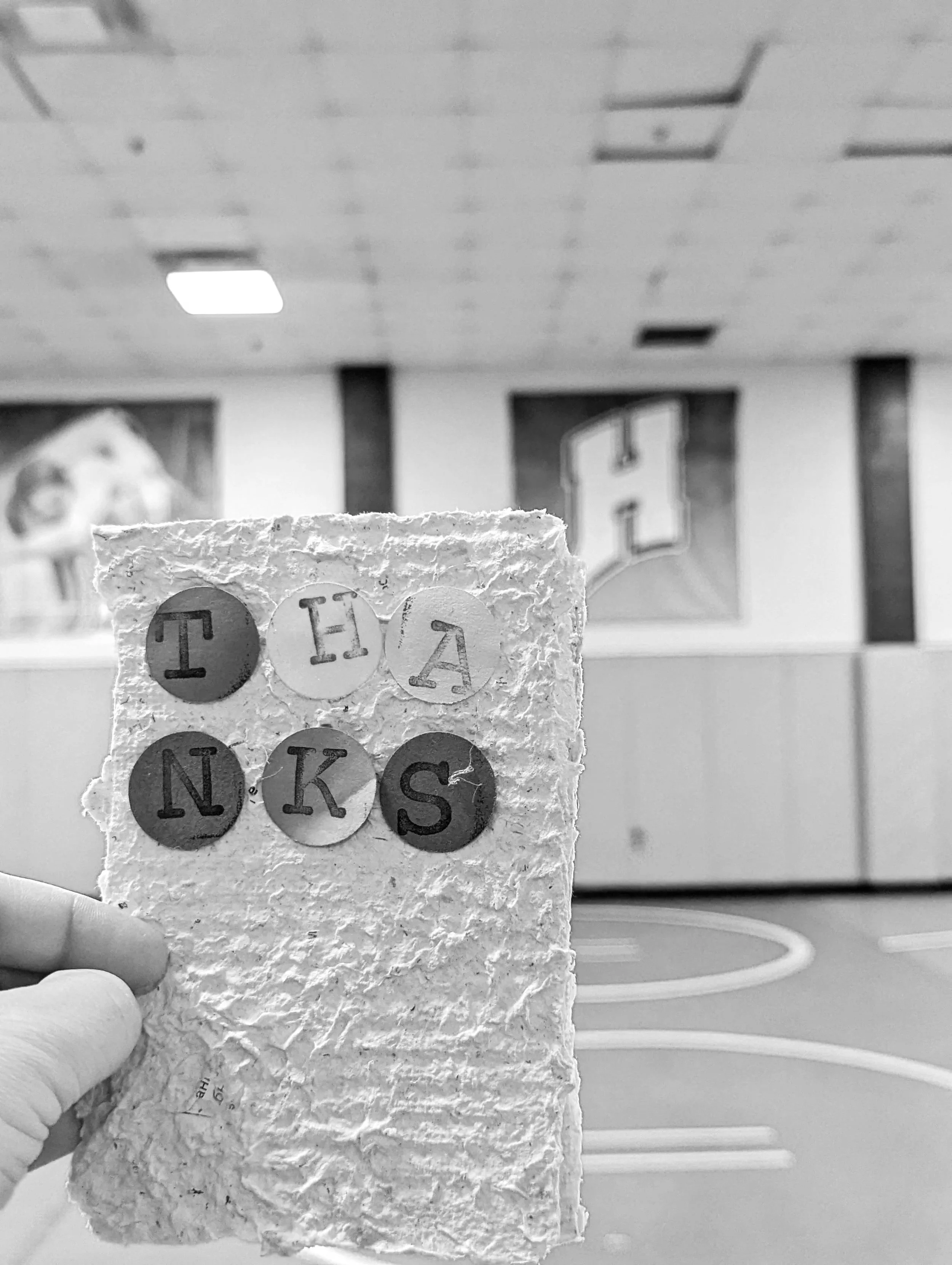 A black and white photo of a hand holding a piece of textured paper with letters on circular stickers, reading "TEAK". The background shows a room with a fluorescent light on the ceiling, a window, and part of a framed picture on the wall.
