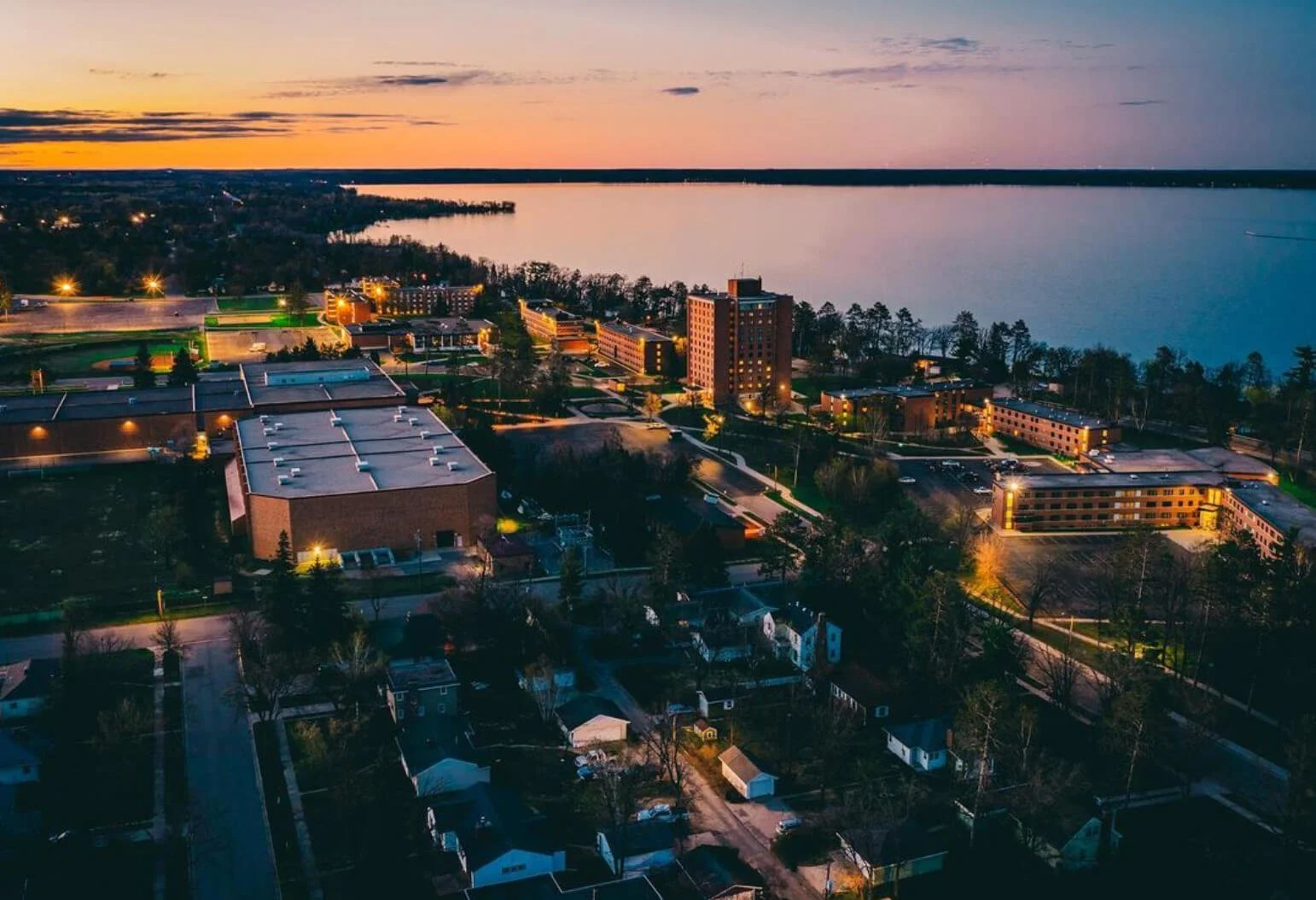 An aerial view of a cityscape at sunset, showing residential houses, a large building, leafy trees, and a body of water in the background.