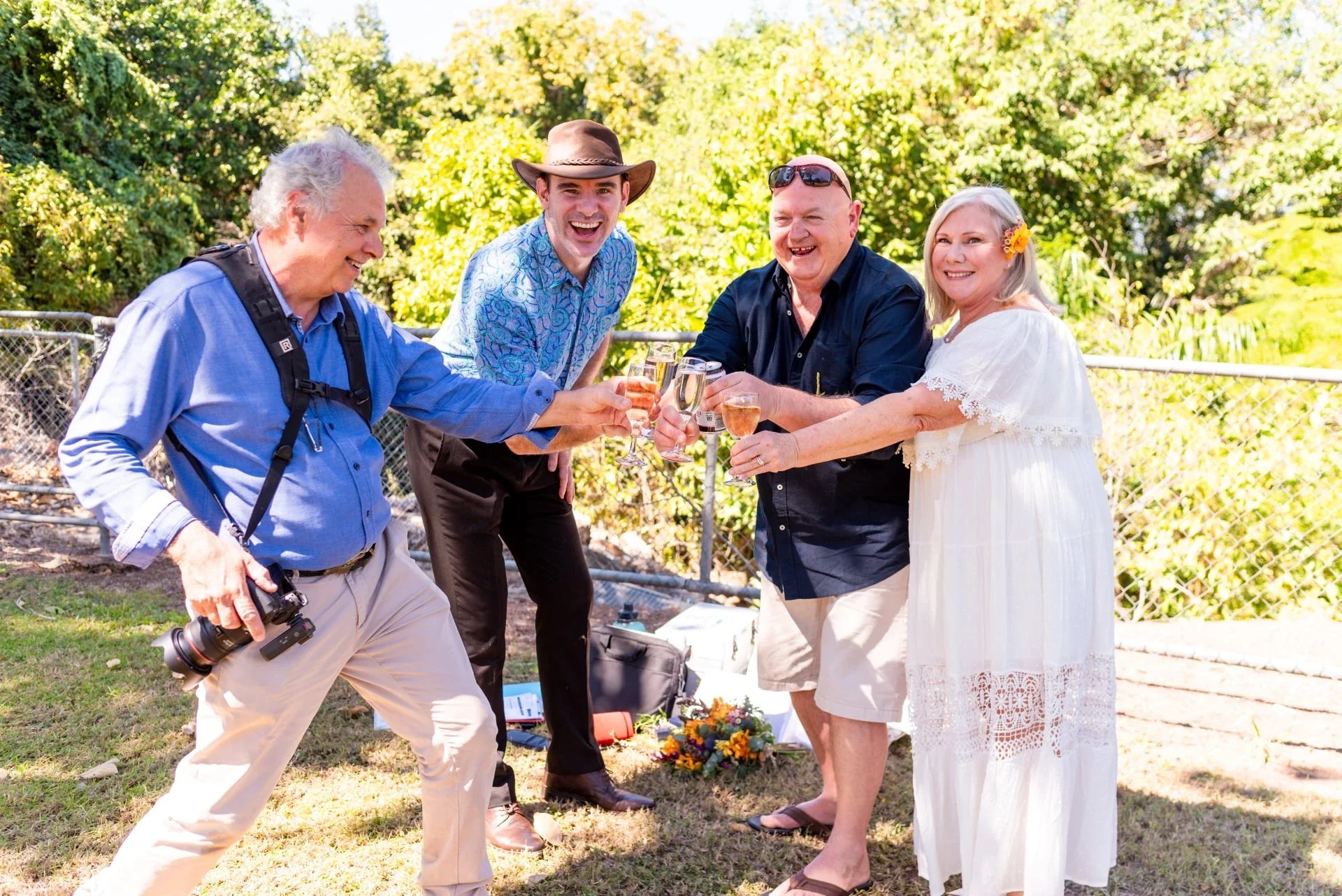 Group of five adults celebrating outdoors with drinks, smiling and toasting, in a sunny garden with trees.
